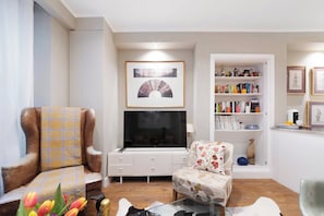 Una sala de estar con un sillón de cuero marrón, un sofá blanco, una mesa de centro de cristal, un televisor de pantalla plana y una estantería llena de libros y objetos decorativos.
