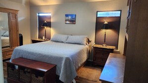 A bedroom with a bed, bedside lamps, a wooden bench, a mirror, and a window with blinds. - Texas Motor Speedway Comfort Zone (Justin)