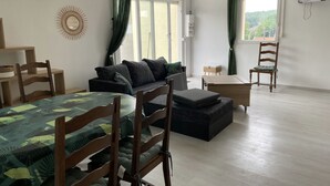 A well-lit room with a sofa, a wooden chair, a small table, and a bed with a green and black patterned bedspread.
