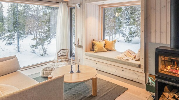 A cozy room with a wooden interior, a fireplace, a seating area with cushions, and a view of a snowy forest through large windows. - 3 bedroom amazing home in Tuddal (Tuddal)