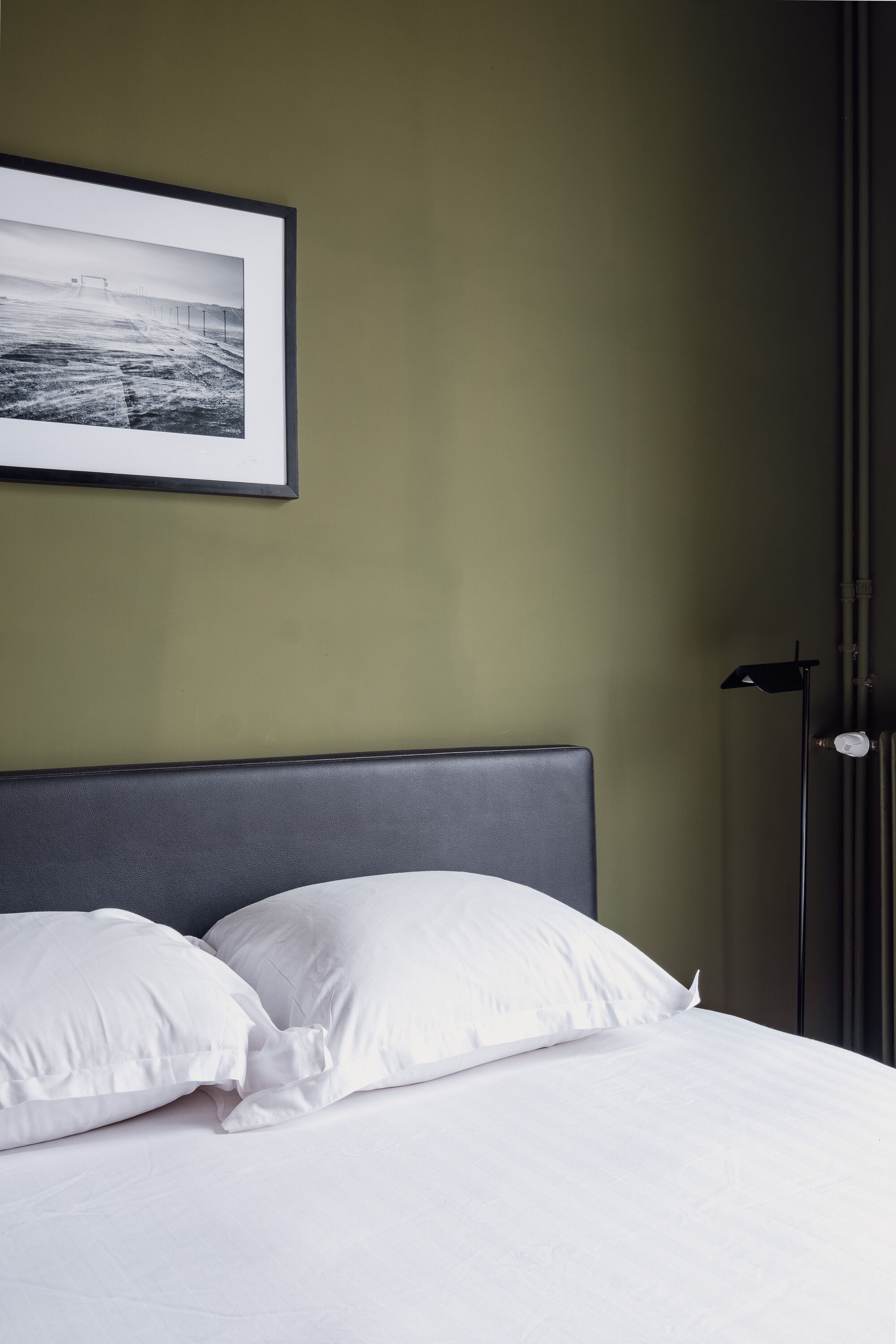 Una cama bien hecha con sábanas y almohadas blancas, un cuadro en la pared y una lámpara de pie junto a la cama.