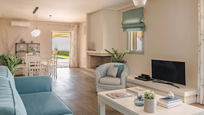 Una sala de estar con televisor, sofá, sillón y chimenea. Hay un comedor con mesa y sillas, y vistas al exterior.