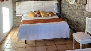 Une chambre avec un mur en pierre, un plafond en bois, un lit avec une literie blanche et une petite table blanche.