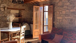 Une pièce chaleureuse avec un mur en pierre, des poutres apparentes en bois au plafond, une table à manger avec des chaises, un canapé et un ordinateur portable posé sur une petite table.
