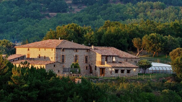 Un bâtiment en pierre, surmonté d’un toit en tuiles, entouré d’une végétation luxuriante et d’arbres.