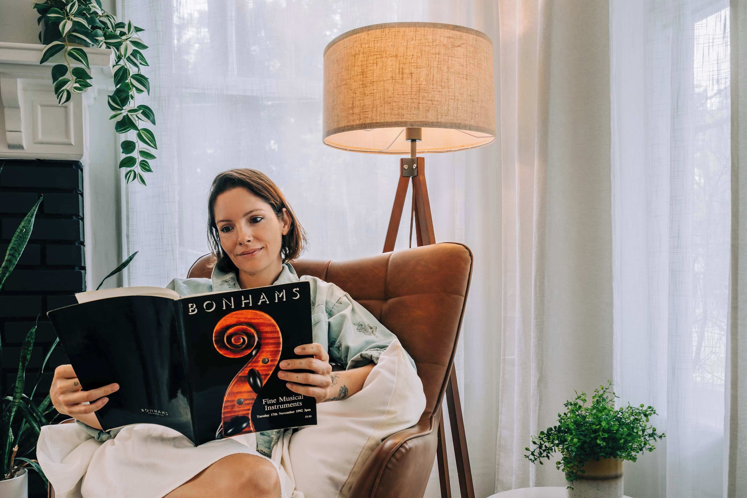 Una persona leyendo un libro titulado "BONHAMS" en una habitación con una lámpara, plantas en macetas y una ventana.