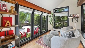 A modern living room with large windows, a bookshelf, and a seating area with patterned cushions. - Retro Razorback - Pickleball-Golf-Spa-UofA (Fayetteville)