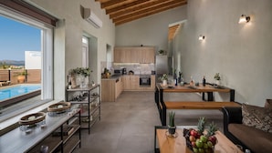 A modern kitchen with wooden cabinets, a dining area with a wooden table, and a pool visible through the window. - Roxanne Luxury Villa, Private Pool & Seaview (Chania)