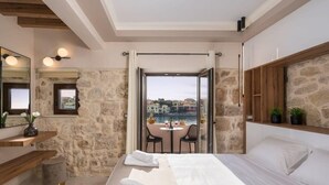 Ein Hotelzimmer mit Steinwand, einem Bett mit Handtüchern, einem kleinen Tisch mit Stühlen und Blick auf eine Stadtlandschaft durch eine Glasdoor.