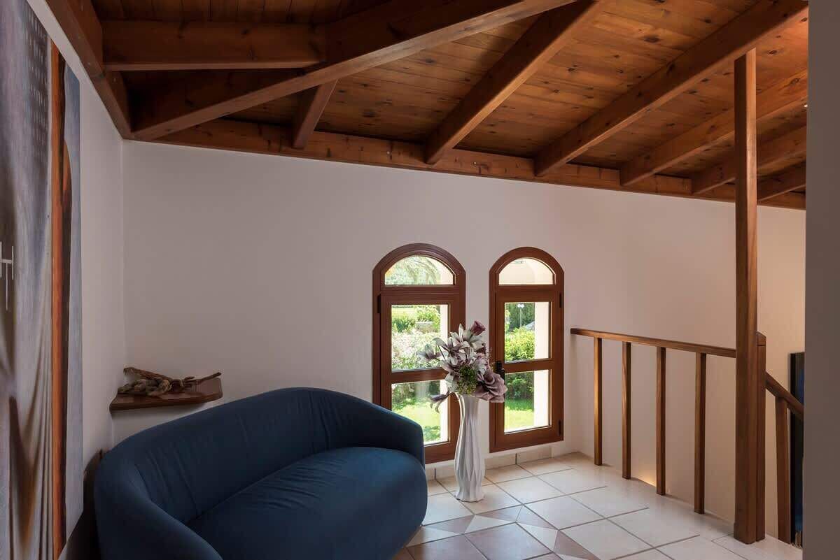 A room with a wooden ceiling, a blue sofa, a white vase with flowers, and two arched windows.