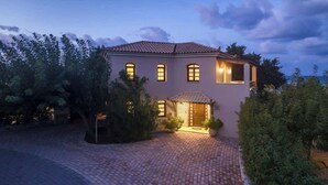 A two-story house with a tiled roof, a brick driveway, and a well-lit entrance. - Argo Luxury Villa (Iason Villas), 3 Bedrooms (Maleme)