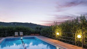A rectangular swimming pool with a clear blue water surface, surrounded by a tiled deck and a metal railing. There are two lounge chairs and a small seating area with a table. The background features a hilly landscape with a sunset sky. - Argo Luxury Villa (Iason Villas), 3 Bedrooms (Maleme)