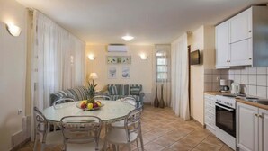 A dining area with a round glass table, chairs, and a fruit bowl. There is a sofa with striped cushions and a lamp. The kitchen area includes white cabinets and a stove. - Argo Luxury Villa (Iason Villas), 3 Bedrooms (Maleme)