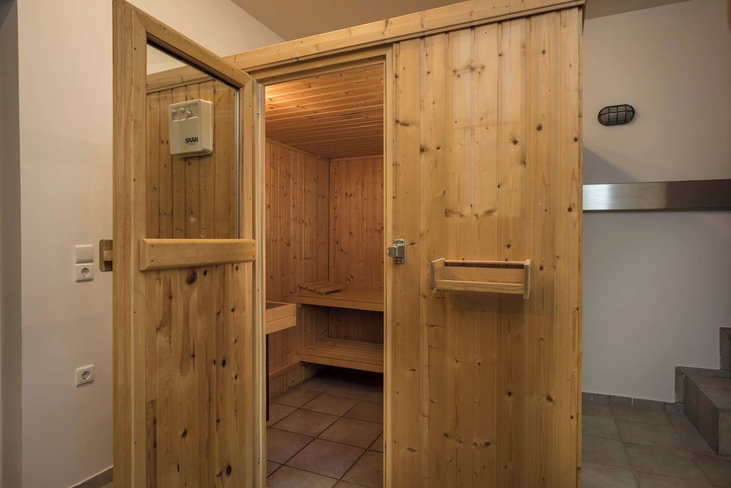 Une cabine de sauna en bois, dotée d’une porte, d’une petite étagère et d’un sol carrelé.