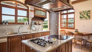A kitchen with granite countertops, wooden cabinets, a built-in stove, and a window overlooking a garden. - Natali Luxury Villa (Iason Villas), 9 Bedrooms (Chania)