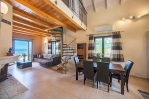 A spacious living area with a wooden ceiling, a dining table with chairs, a staircase, and a view through a large window.