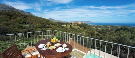 Una mesa de madera dispuesta con platos, cubiertos y un frutero en un balcón con vistas a un paisaje montañoso y una piscina.