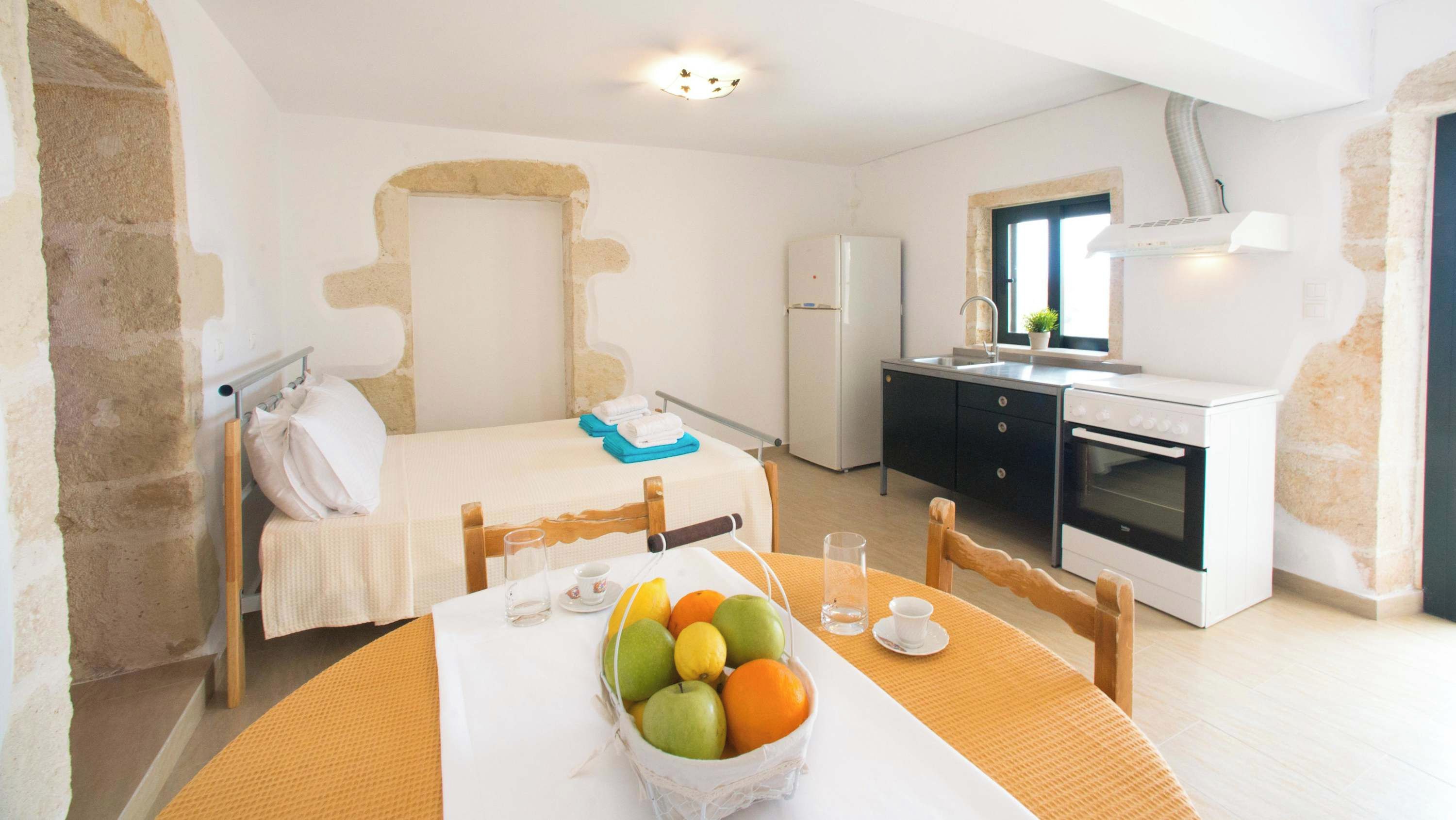A compact hotel room with a bed, a small dining table with fruit, a kitchenette, and a bathroom.