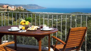 Une table en bois, dressée pour le petit-déjeuner, avec deux chaises, et une vue magnifique sur la mer et les bâtiments.