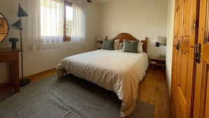 Un dormitorio con cama, mesita de noche, lámpara, armario de madera y ventana con cortinas.