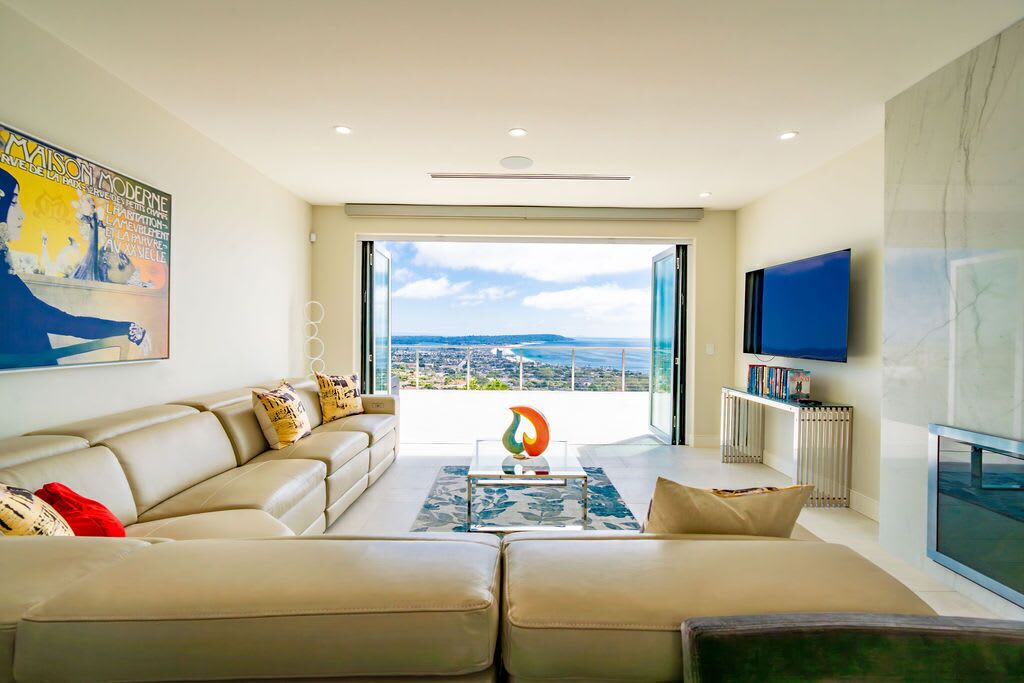 A modern living room with a large sofa, a coffee table, and a television. There is a large window providing a view of the city.