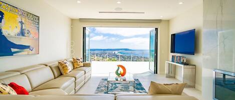 A modern living room with a large sofa, a coffee table, and a television. There is a large window providing a view of the city.