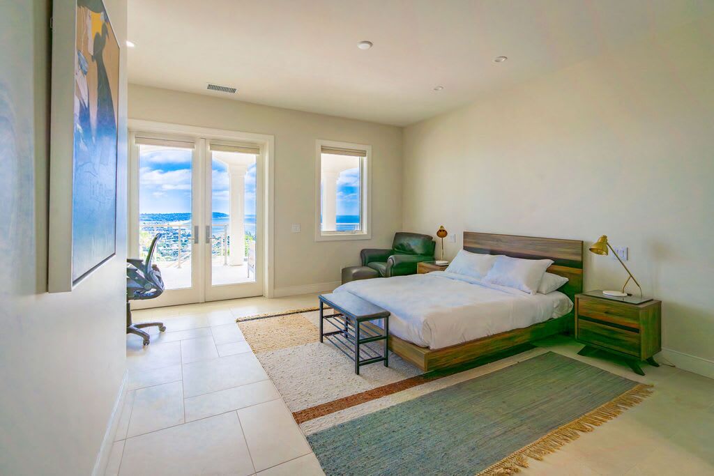 A bedroom with a bed, a desk, a chair, and a view of the ocean through large windows.
