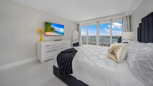 A modern bedroom with a large bed, a flat-screen TV, a dresser, and a view of the ocean through large windows.