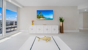 A modern living room with a large window, a flat-screen TV mounted on the wall, a white console table, and a decorative fish sculpture on the floor.