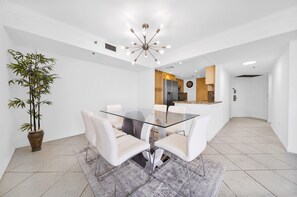 A modern dining area with a glass table and white chairs, a bamboo plant, and a kitchen in the background. - Open Living Bay Views | 36th Floor | StayCozy (Miami)