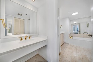 A modern bathroom with a large mirror, a white sink with gold fixtures, and a bathtub. - Open Living Bay Views | 36th Floor | StayCozy (Miami)