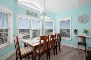 Un comedor con una mesa y sillas de madera, ventanas amplias con persianas, un reloj y un letrero de bienvenida.