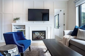 A living room with a fireplace, a flat-screen TV mounted above it, a blue armchair, a grey sofa, and a coffee table.