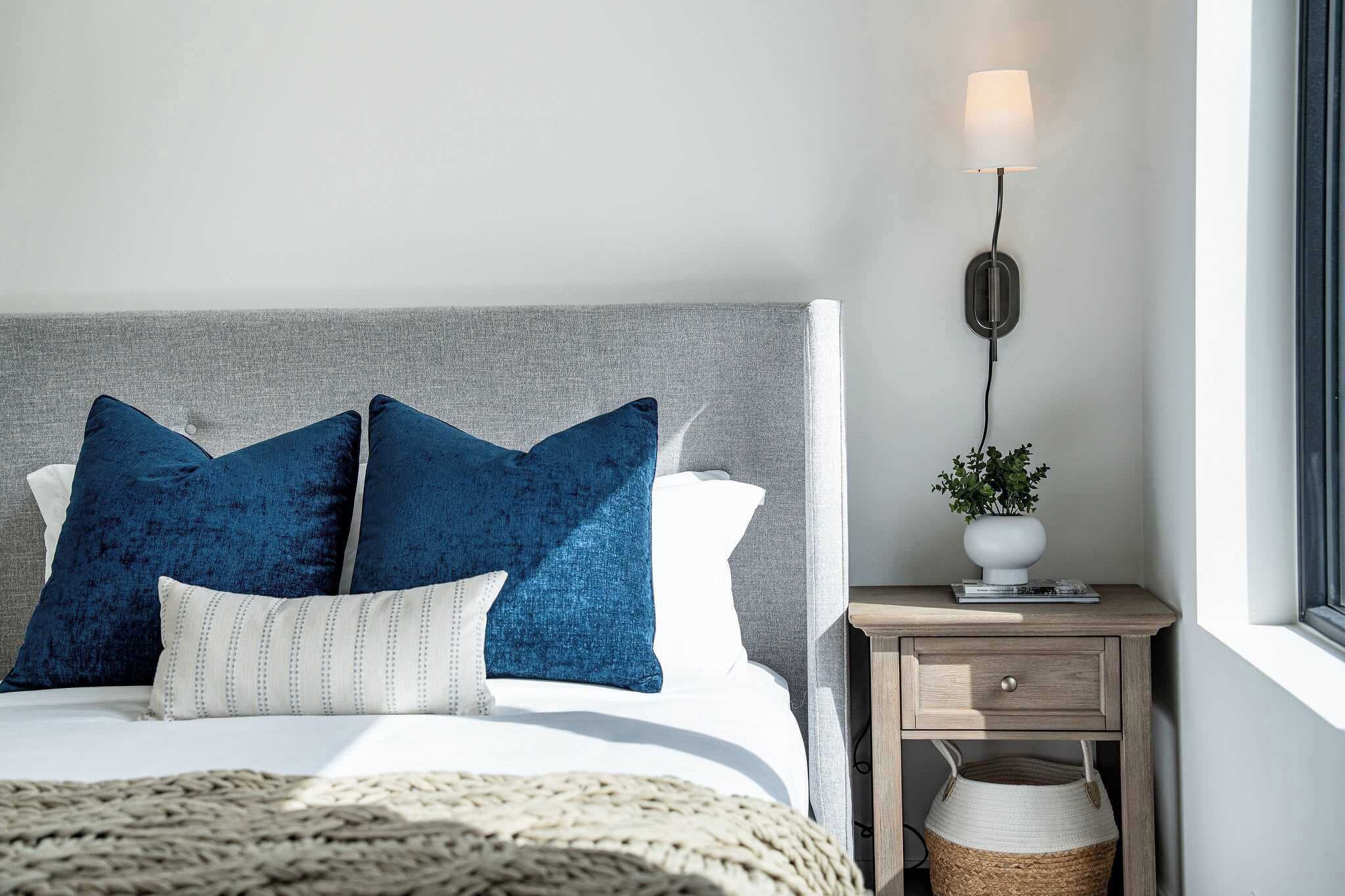 A bed with blue pillows, a bedside lamp, a small table with a plant, and a window.
