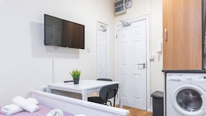 A compact hotel room with a bed, a small table with a plant, a TV mounted on the wall, a washing machine, and a door with a coat rack. - Cosy citycentre flat with TV & Wi-Fi included (Dublin 1)