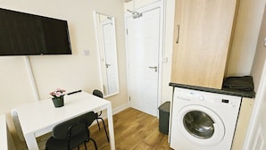 A compact kitchen with a washing machine, a dining table with chairs, and a TV mounted on the wall. - Cosy citycentre flat with TV & Wi-Fi included (Dublin 1)