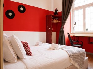 Een slaapkamer met een bed, kussens, een rode garderobe, een bureau en een raam.
