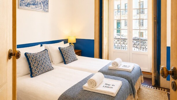 Ein Hotelzimmer mit zwei Betten, einem Gemälde an der Wand, einer Tür, die auf einen Balkon führt, und Handtüchern mit dem Schriftzug 'Stay in Lisbon'.