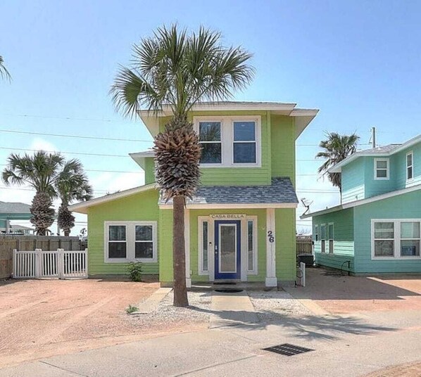 Una casa de dos pisos con una puerta azul, con el número 26, y una palmera enfrente.