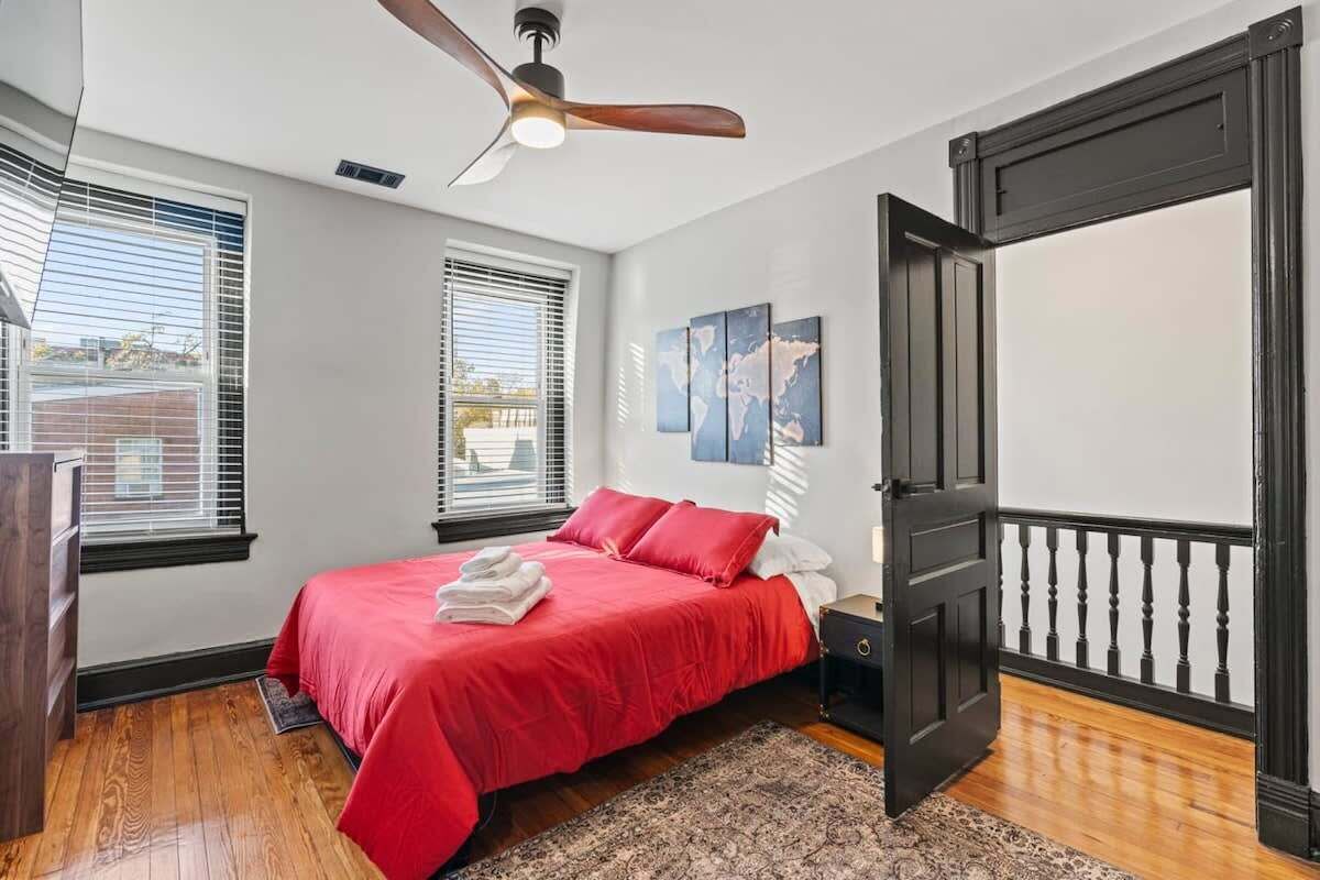Ein Schlafzimmer mit roter Bettdecke, Deckenventilator, einer Weltkarte als Wandbild und Blick ins Freie durch das Fenster.