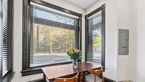 Un comedor con una mesa de madera y dos sillas, un florero con flores, y vistas a través de ventanas con marco negro.