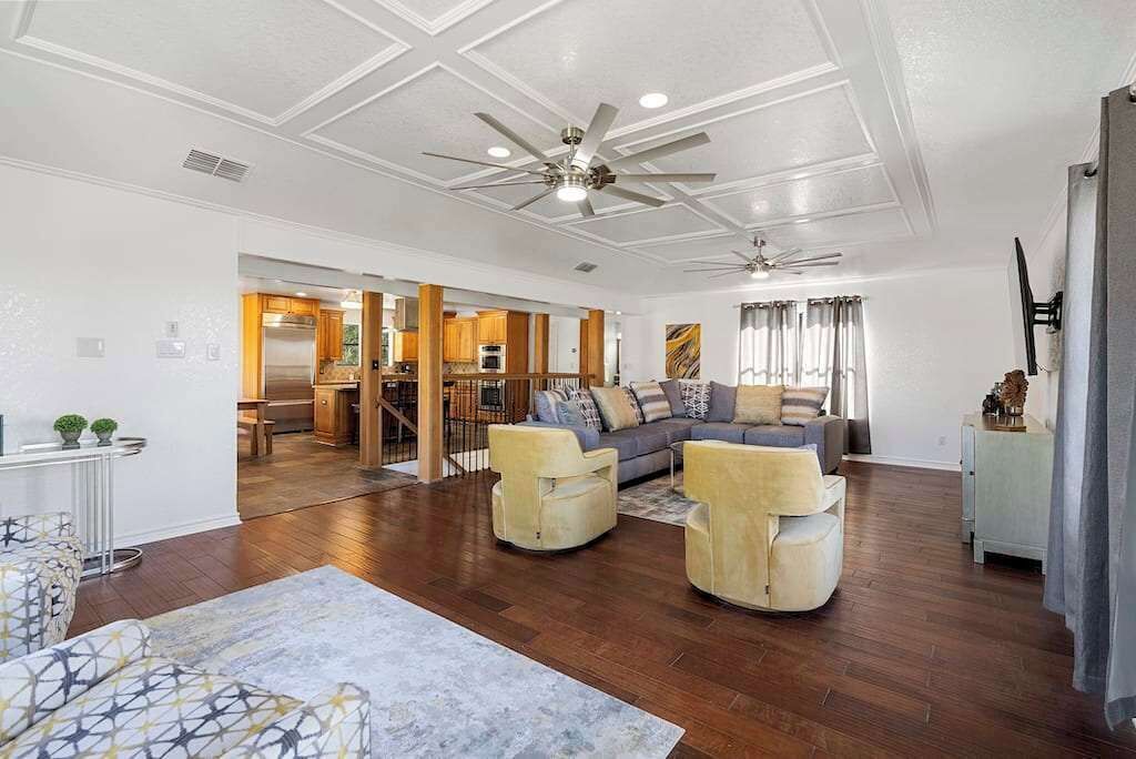 A spacious living room with a large sofa, two armchairs, a coffee table, and a ceiling fan. There is a kitchen visible in the background.