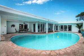 A swimming pool with a covered patio area, a dining table, and chairs.