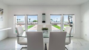 A modern dining area with a glass table and white chairs, large windows with a view of a landscaped area. - Sleep 29 - Oceanfront Retreat 2 Homes for 29-Guests! (Corpus Christi)