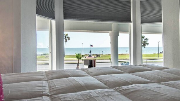 A hotel room with a large bed, a view of the beach, and a flagpole with an American flag. - Sleep 29 - Oceanfront Retreat 2 Homes for 29-Guests! (Corpus Christi)