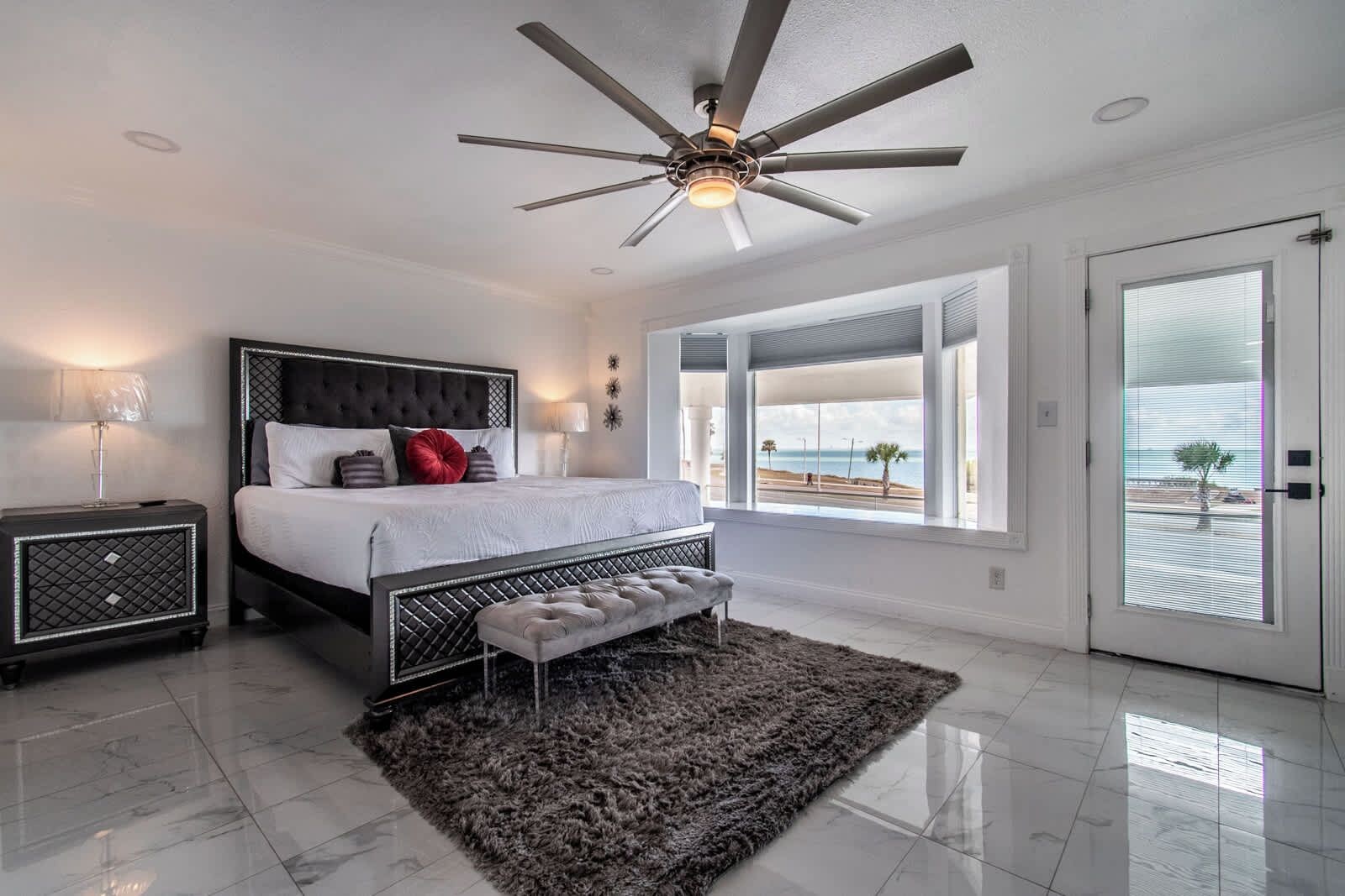 Un dormitorio moderno con cama, mesitas de noche, ventilador de techo y vista a la playa.