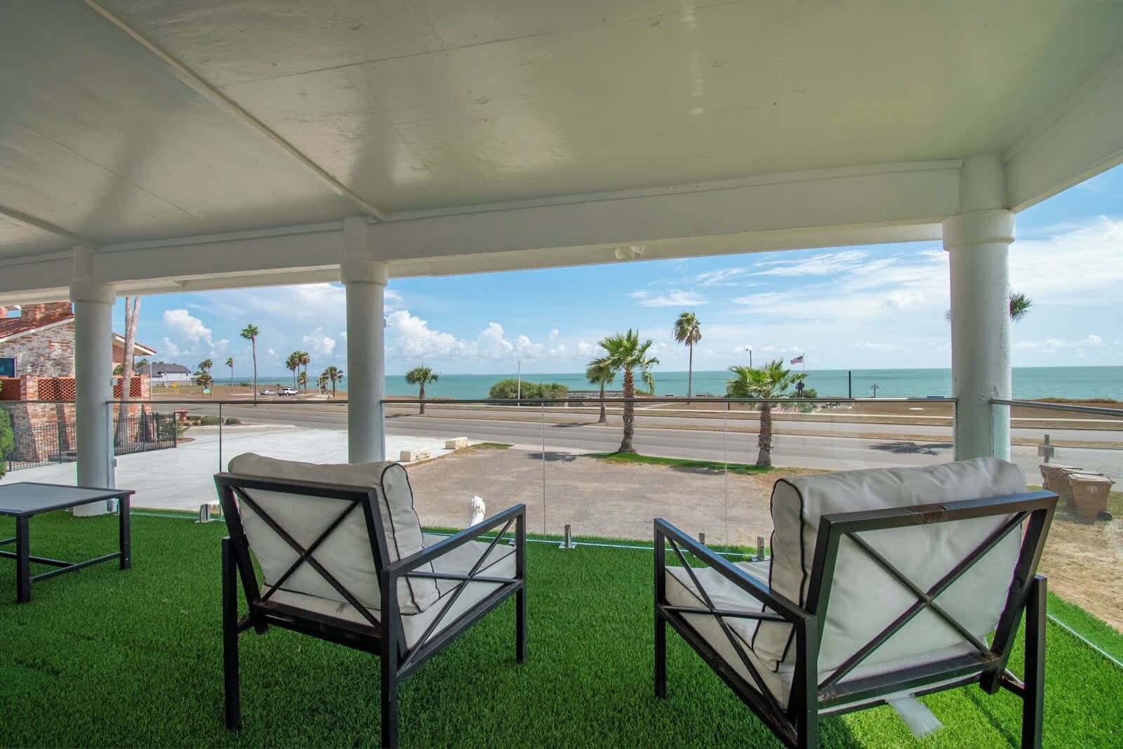 Un espacio de estar techado al aire libre con dos sillas y una mesa, con vistas a una carretera y una playa con palmeras.