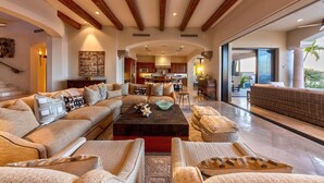 A spacious living room with a large sectional sofa, a coffee table, and a wicker chair. A kitchen is visible in the background, and there is a large sliding glass door leading to an outdoor area. - 5BR Luxury Home in Villas del Mar (Los Cabos)