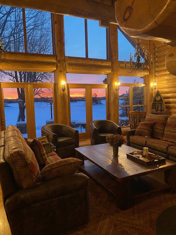 A cozy log cabin interior with leather furniture, a wooden coffee table, and large windows offering a view of a snowy landscape.