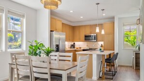 Une cuisine moderne avec des meubles en bois, des appareils électroménagers en acier inoxydable, un îlot central, des luminaires suspendus et un coin repas avec des chaises blanches.